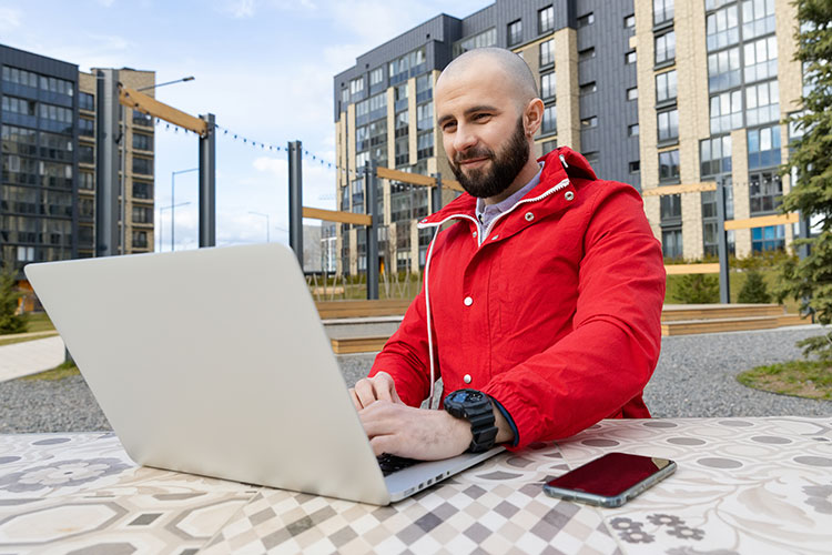 Man in red jacket using laptop outdoors.